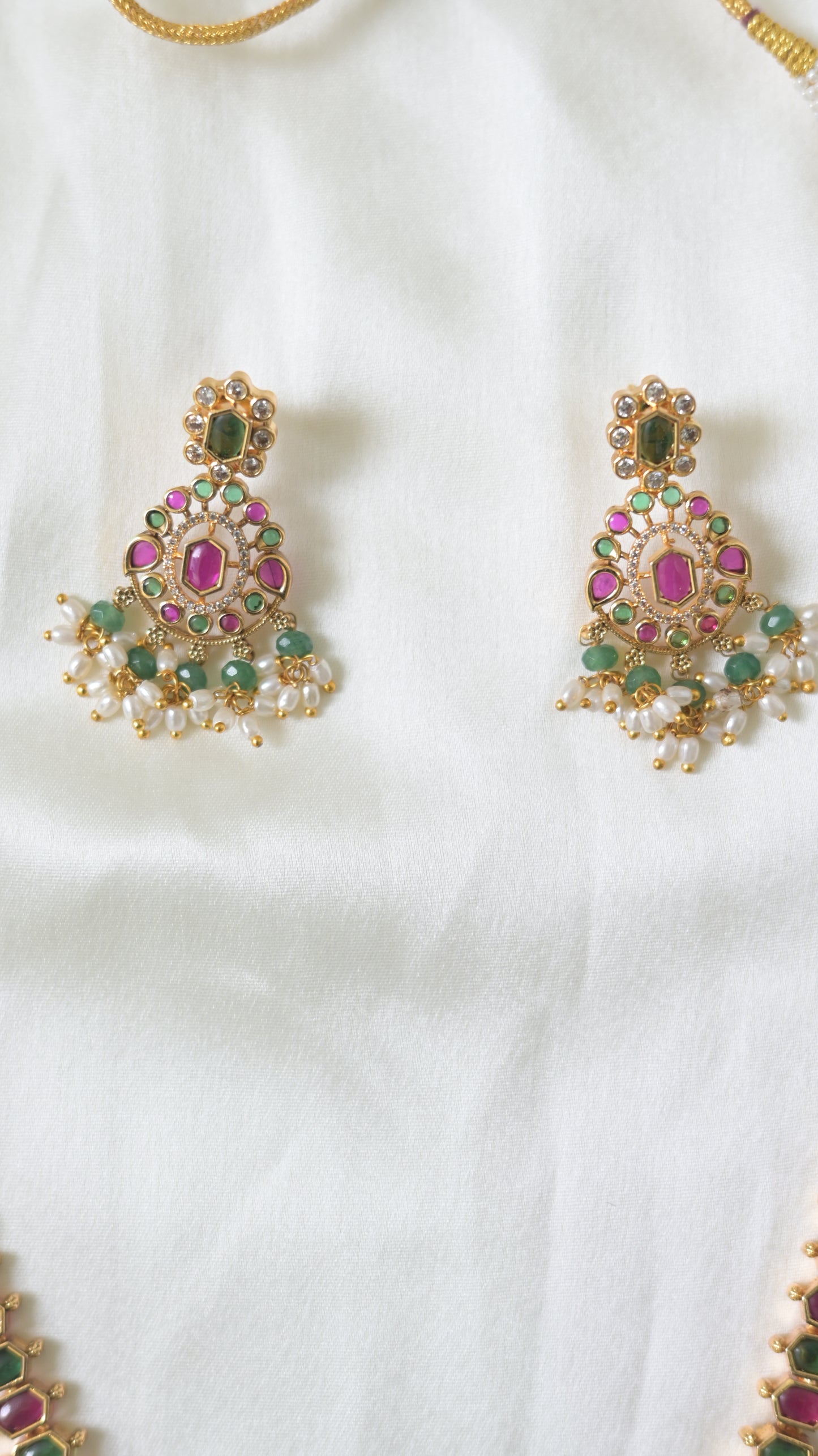 Viyaraa Jewels traditional gold-plated long haram necklace and matching earrings set. The jewelry features a continuous pattern of alternating ruby-pink and emerald-green stones bordered by clear stones, culminating in chandelier-style pendants with white pearl and green bead drops, displayed against a white background.