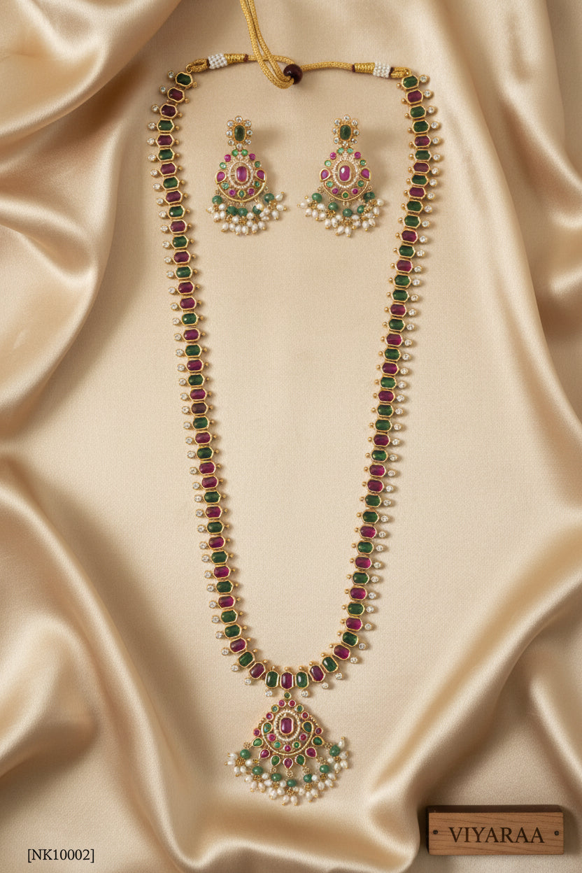 Viyaraa Jewels traditional gold-plated long haram necklace and matching earrings set. The jewelry features a continuous pattern of alternating ruby-pink and emerald-green stones bordered by clear stones, culminating in chandelier-style pendants with white pearl and green bead drops, displayed against a white background.