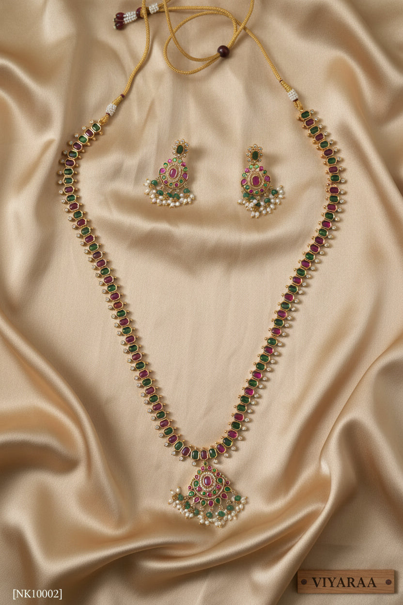 Viyaraa Jewels traditional gold-plated long haram necklace and matching earrings set. The jewelry features a continuous pattern of alternating ruby-pink and emerald-green stones bordered by clear stones, culminating in chandelier-style pendants with white pearl and green bead drops, displayed against a white background.