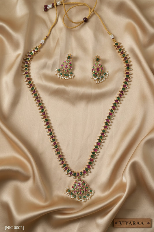 Viyaraa Jewels traditional gold-plated long haram necklace and matching earrings set. The jewelry features a continuous pattern of alternating ruby-pink and emerald-green stones bordered by clear stones, culminating in chandelier-style pendants with white pearl and green bead drops, displayed against a white background.